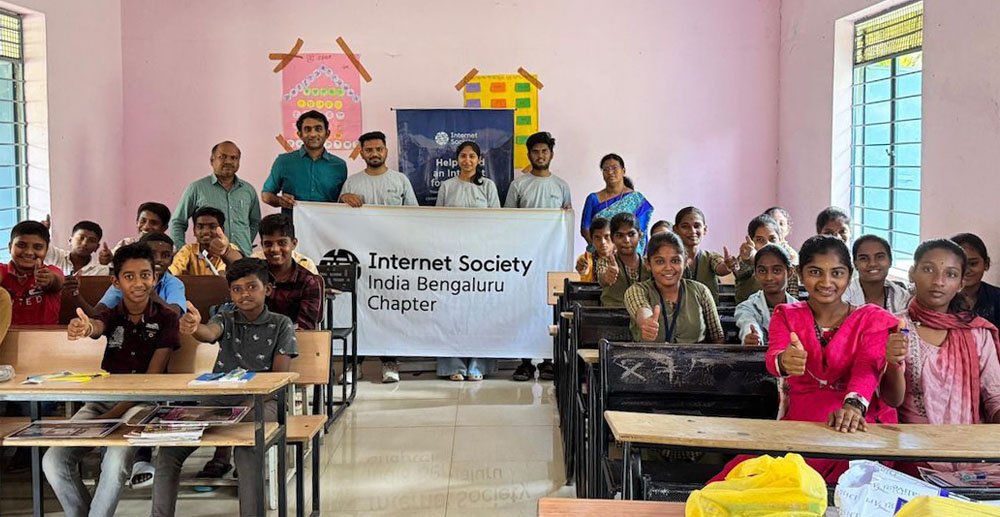 A group of people sit in a classroom and make a thumbs up gesture. People in the back of the room hold a sign that says Internet Society India Bengaluru Chapter